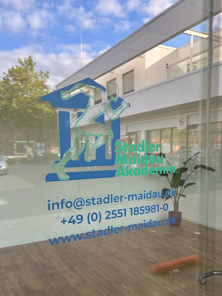 Blick durch die Glasfront der Stadler Maidau Akademie in Rheine: auf der Scheibe das blau-grüne Logo mit E-Mail-Adresse und Telefonnummer, dahinter der Schulungsraum