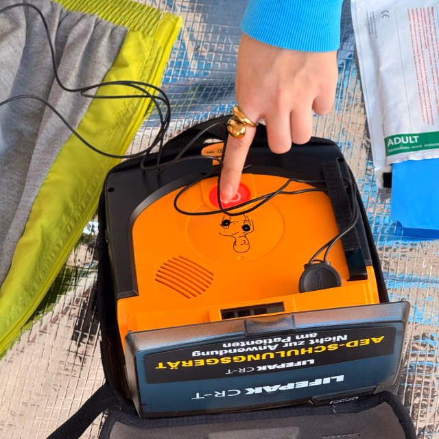 Ein Finger drückt die Schocktaste eines Übungs-Defibrillators, daneben liegt die Übungspuppe mit aufgeklebten Elektroden
