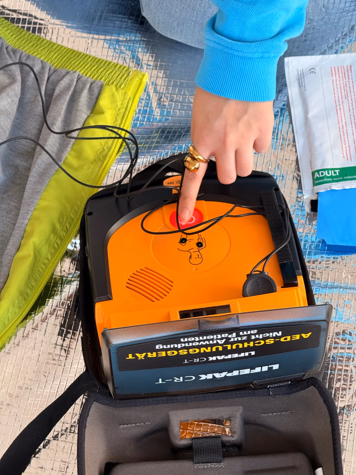Ein Finger drückt die rot leuchtende Schocktaste eines Übungs-Defibrillators, daneben liegt die Übungspuppe mit aufgeklebten Elektroden, Defibrillator-Training im Erste-Hilfe-Kurs