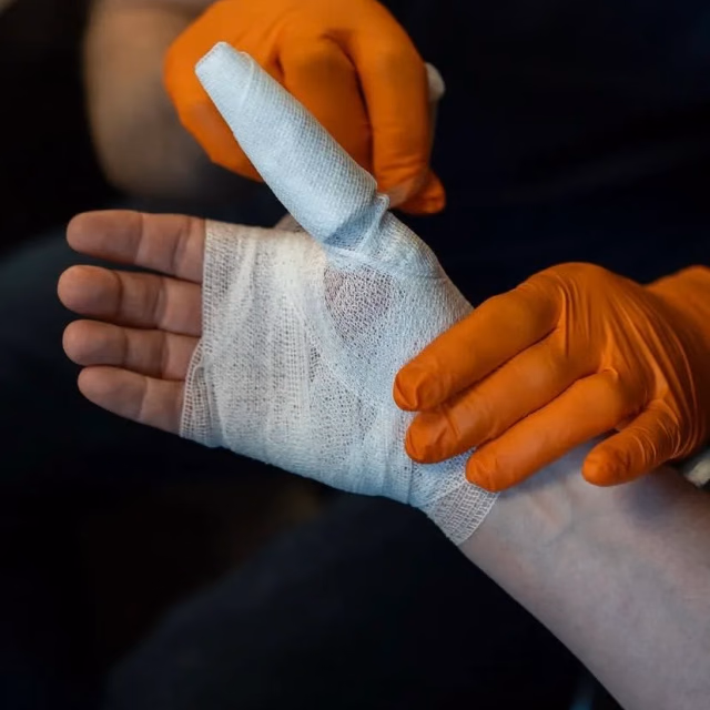 Hand mit frisch angelegtem Mullverband, gehalten von einer Helferin in Einmalhandschuhen