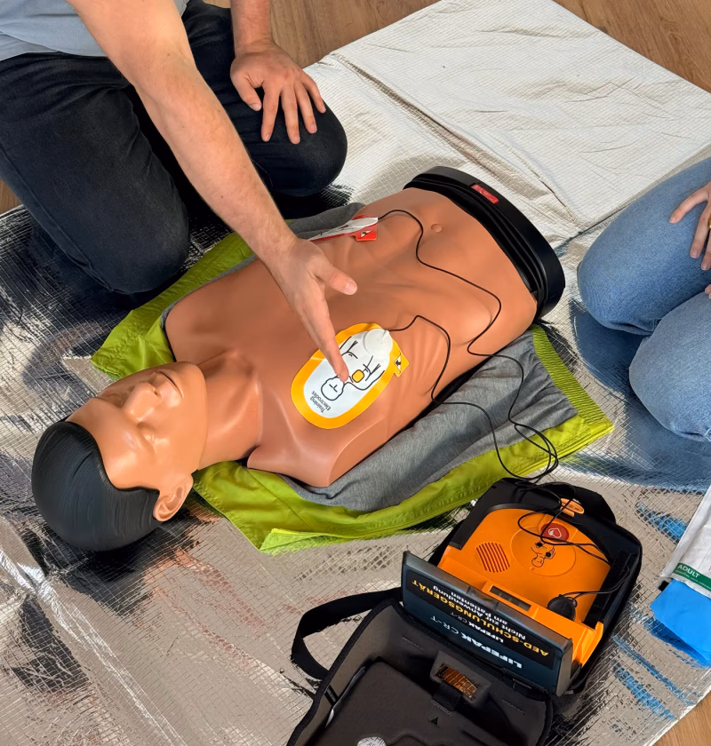 Ausbilder zeigt an der Übungspuppe, wie die Elektrode des Übungs-Defibrillators auf dem Brustkorb sitzt, daneben der geöffnete AED und eine Teilnehmerin auf der Matte
