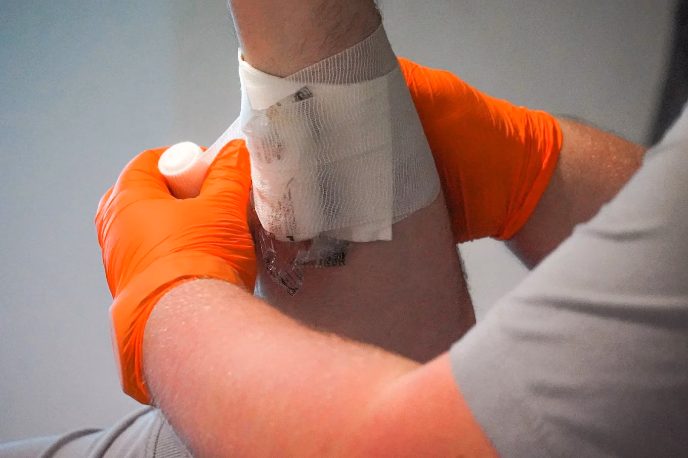 Hände in orangefarbenen Einmalhandschuhen wickeln einen weißen Mullverband um den Unterarm eines Kursteilnehmers