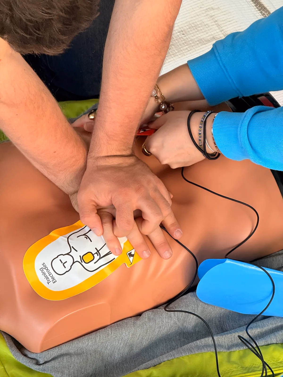 Zwei Hände liegen übereinander auf dem Brustkorb einer Übungspuppe, daneben klebt die Elektrode eines Übungs-Defibrillators, Herzdruckmassage mit angeschlossenem AED