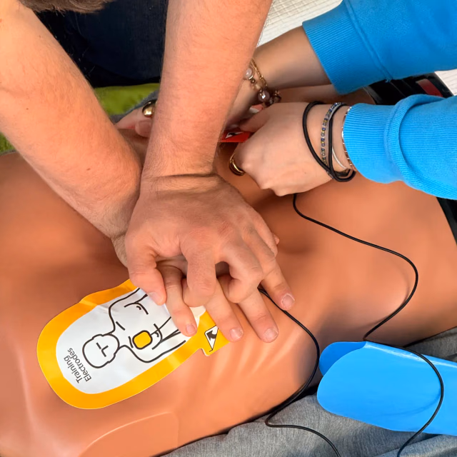 Zwei Hände liegen übereinander auf dem Brustkorb einer Übungspuppe, daneben klebt die Elektrode eines Übungs-Defibrillators