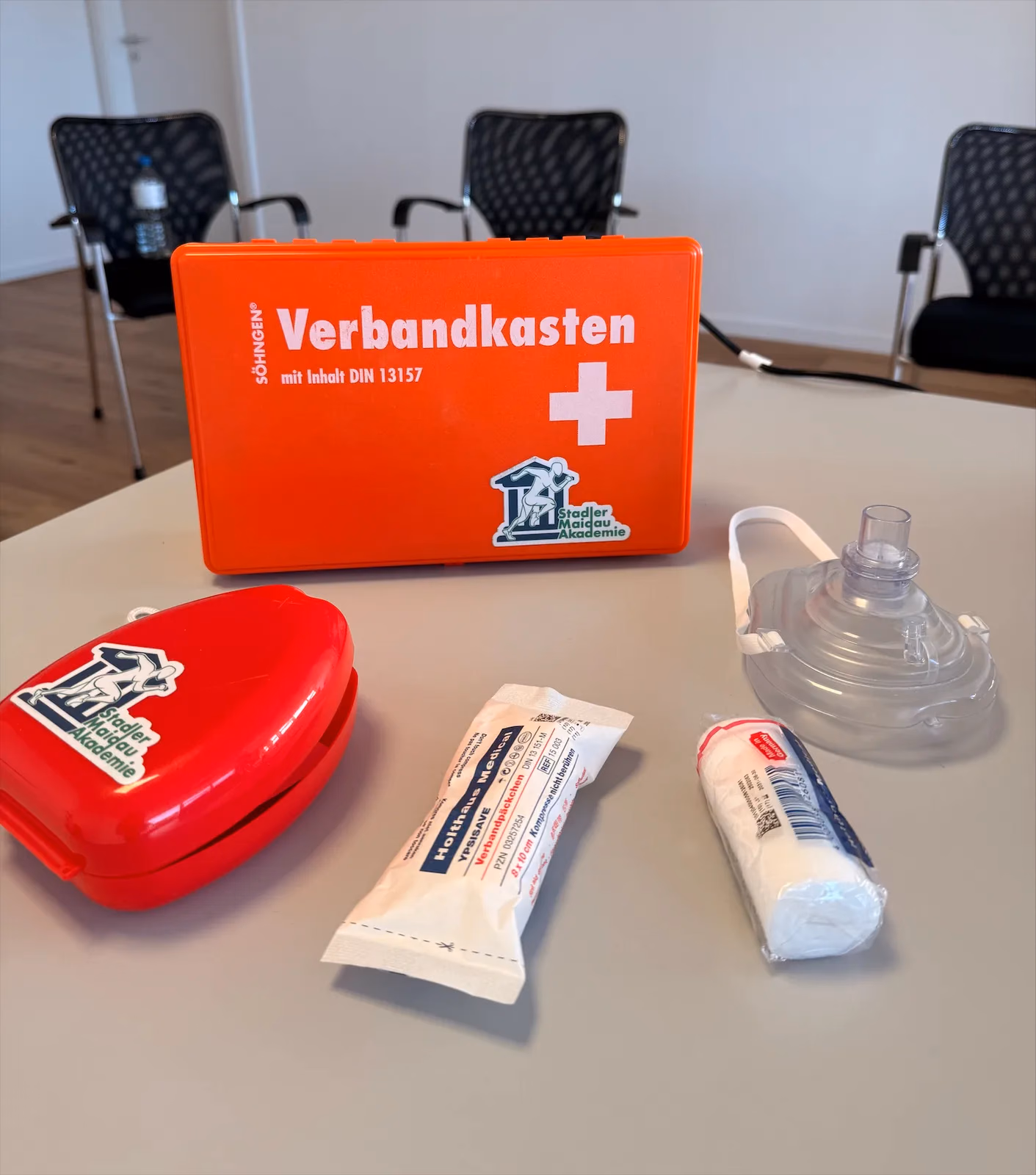 Verbandkasten nach DIN 13157, Erste-Hilfe-Tasche der Stadler Maidau Akademie, Verbandpäckchen und Beatmungsmaske auf einem Tisch im Schulungsraum